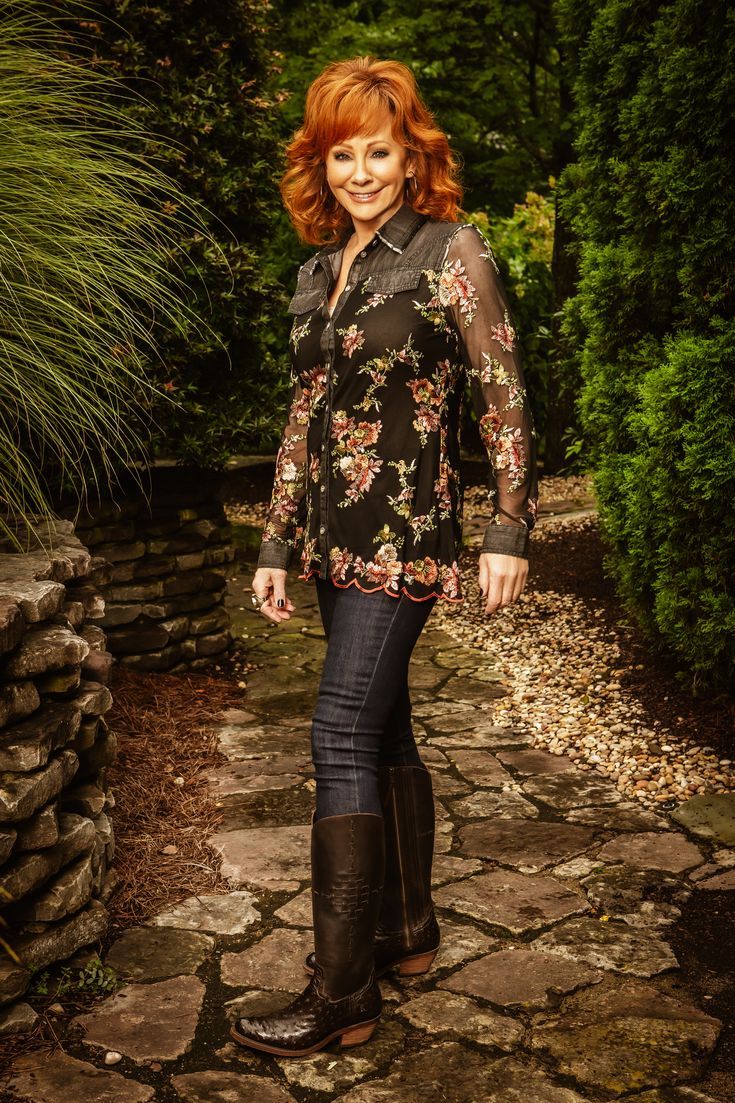 Phần này chứa: Reba McEntire stands in a garden pathway wearing a pair of Reba by Justin boots. She is wearing a black floral top, jeans, and cowboy boots.